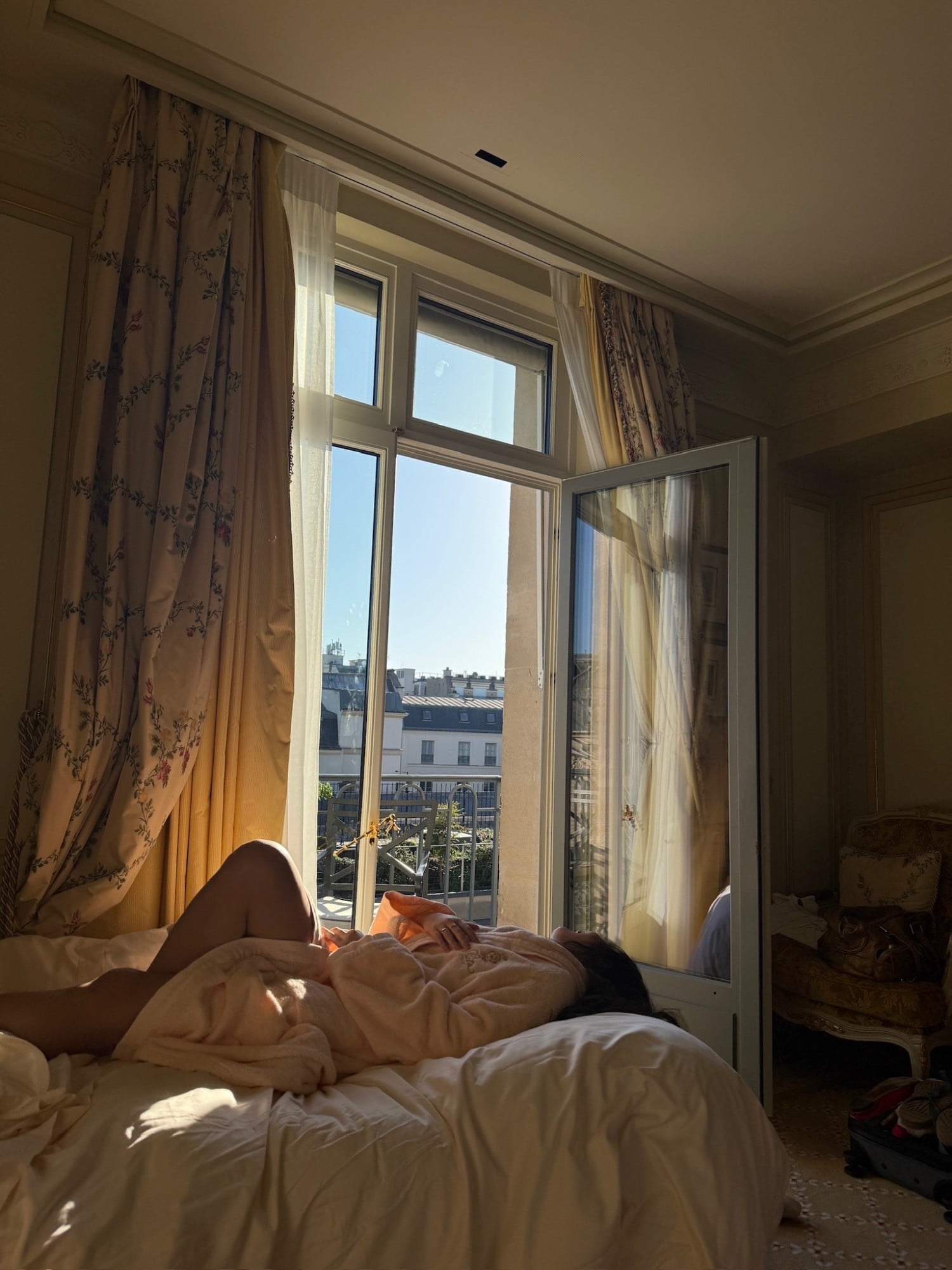 A woman in a robe lying on a bed by an open window in the morning sun